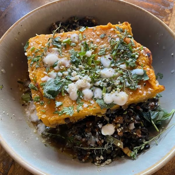 A healthy Strive Foods dish with a golden-brown square of polenta, topped with herbs and white sauce, over a dark lentil mixture.