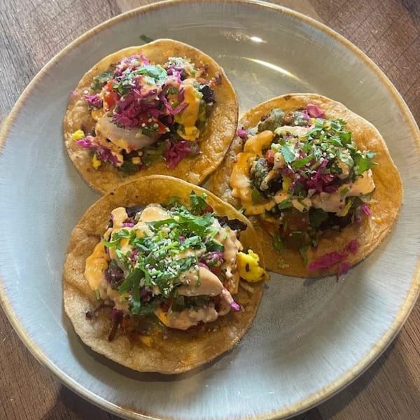 Three healthy tacos from Strive Foods, topped with colorful ingredients and sauce on a light blue plate.